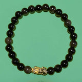 Vue de dessus du Bracelet en perles d'obsidienne noir dorée avec un fond légèrement vert. Une petite figurine représentant un PIXUI fait partir du bracelet.